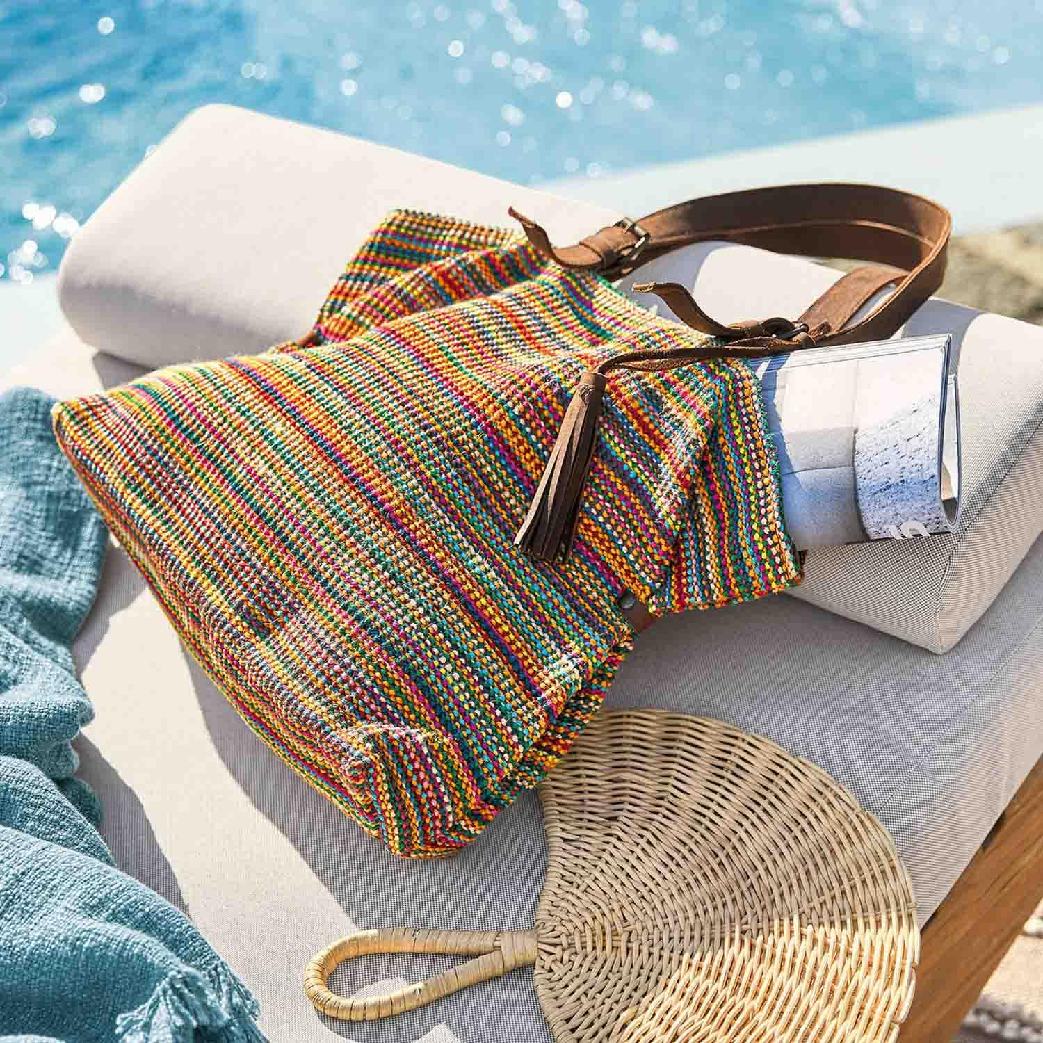 Bolso tejido multicolor con asa de cuero marrón sobre cojín claro junto a la piscina