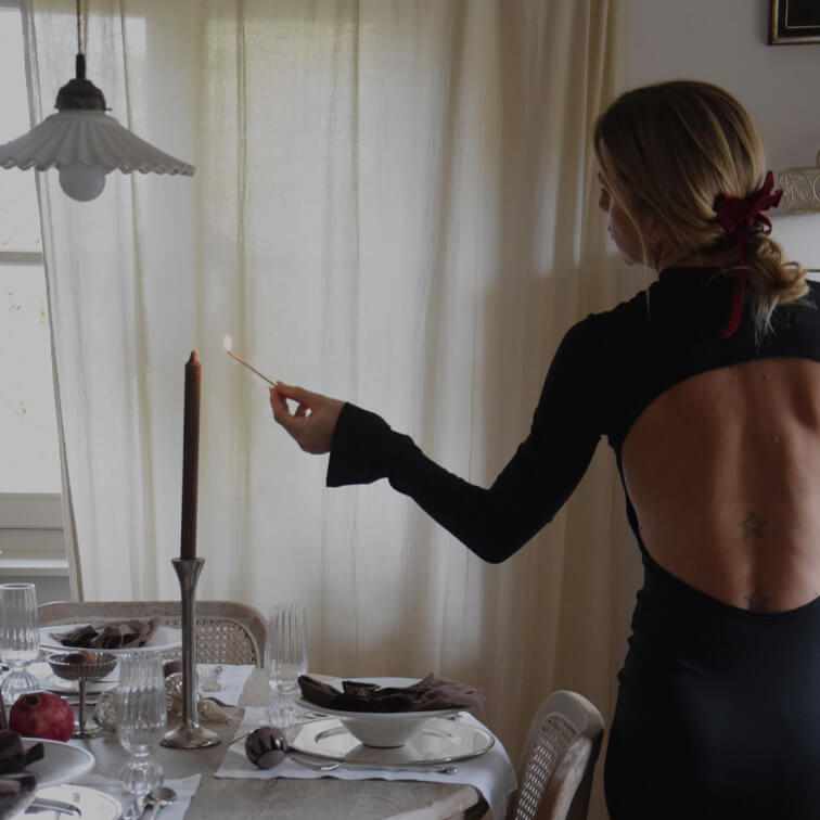 Mujer con vestido negro de espalda descubierta enciende una vela en una mesa elegantemente puesta en el comedor