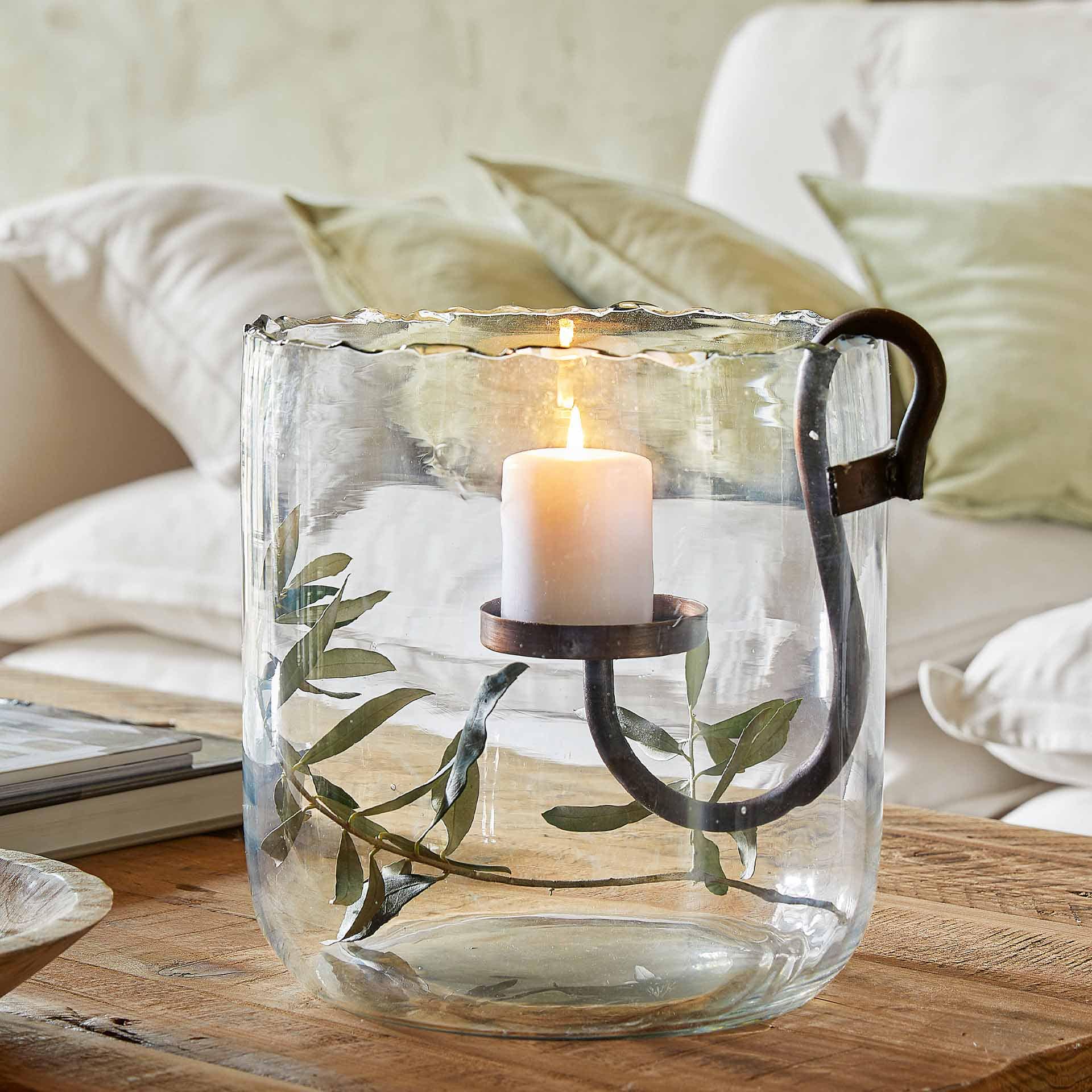 Portavelas de vidrio con asa de metal y vela blanca sobre mesa de madera con hojas verdes
