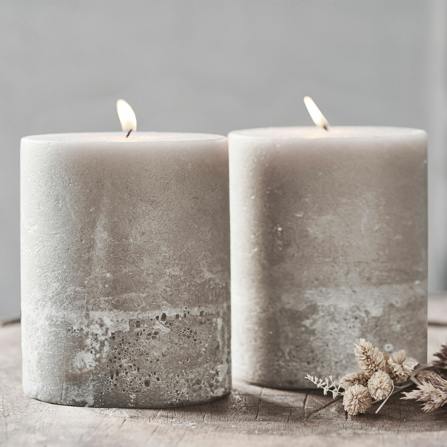 Set de 2 velas Oviedo Dos velas grises de cera con textura rústica sobre mesa de madera con flores secas