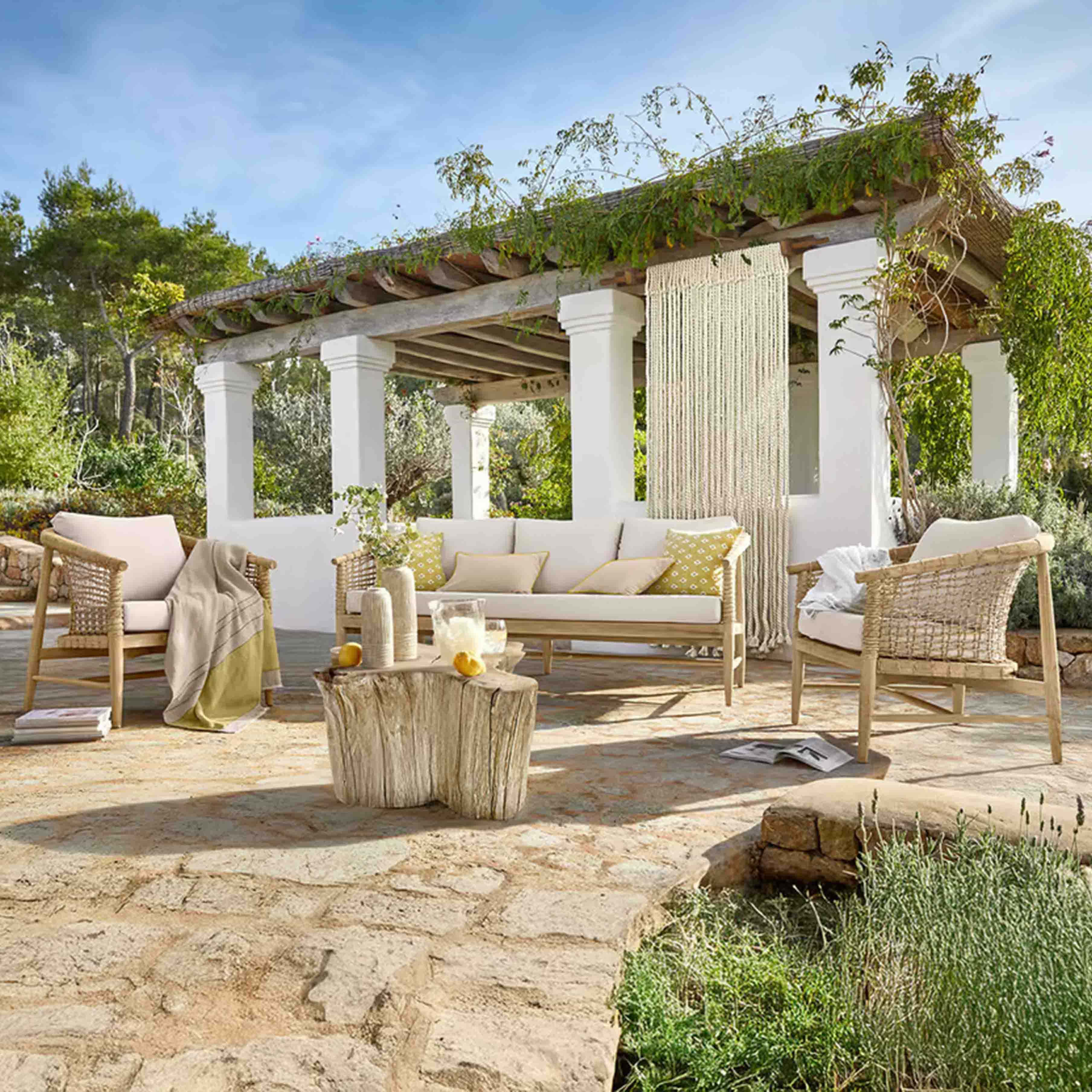 Conjunto de muebles de jardín de madera clara con cojines beige en patio de piedra bajo una pérgola con plantas