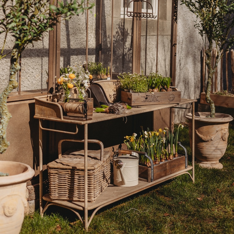 Mesa de jardín de metal con cajas de madera y flores en macetas sobre césped junto a un invernadero