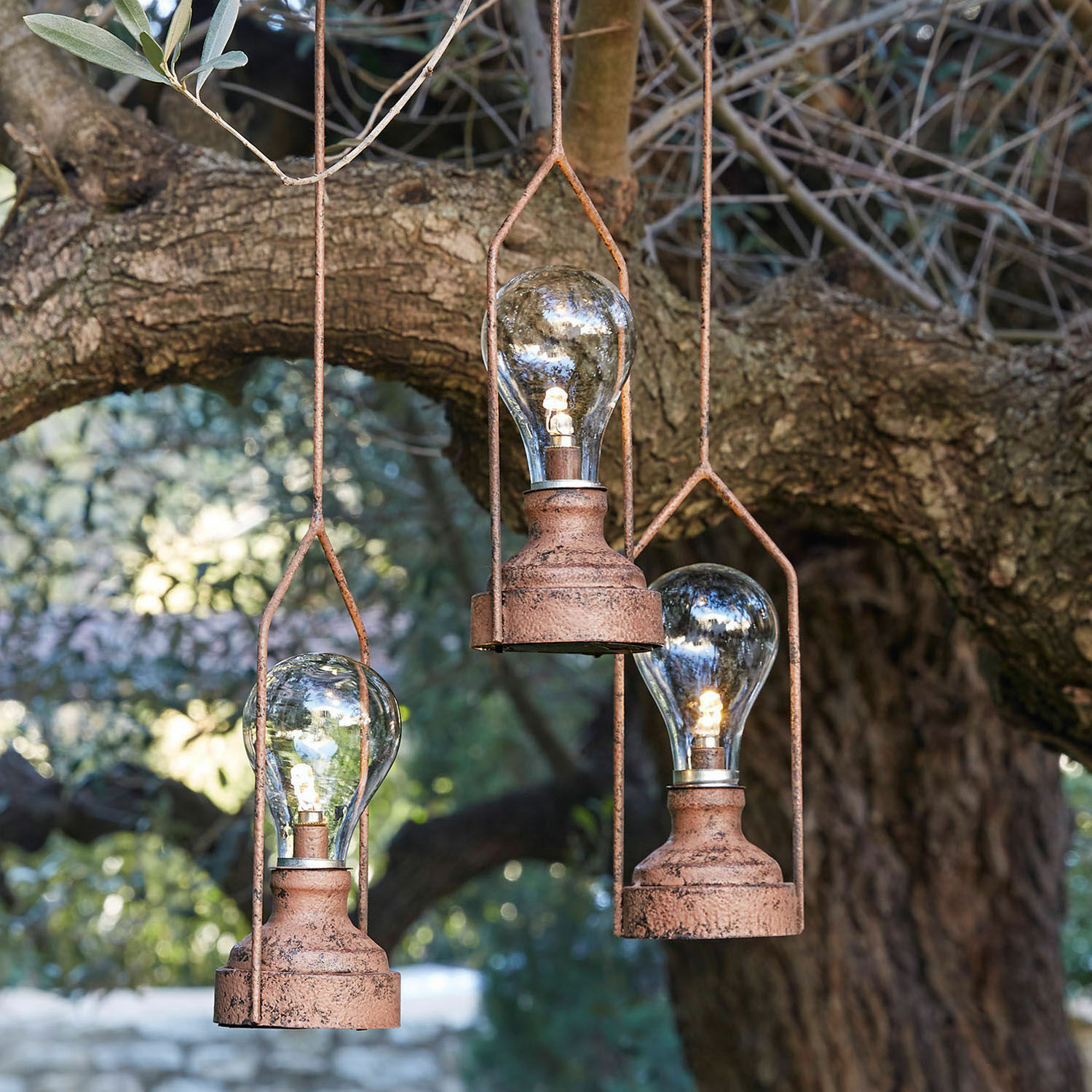 Tres lámparas colgantes de metal oxidado con bombillas LED frente a un árbol, iluminación decorativa de jardín
