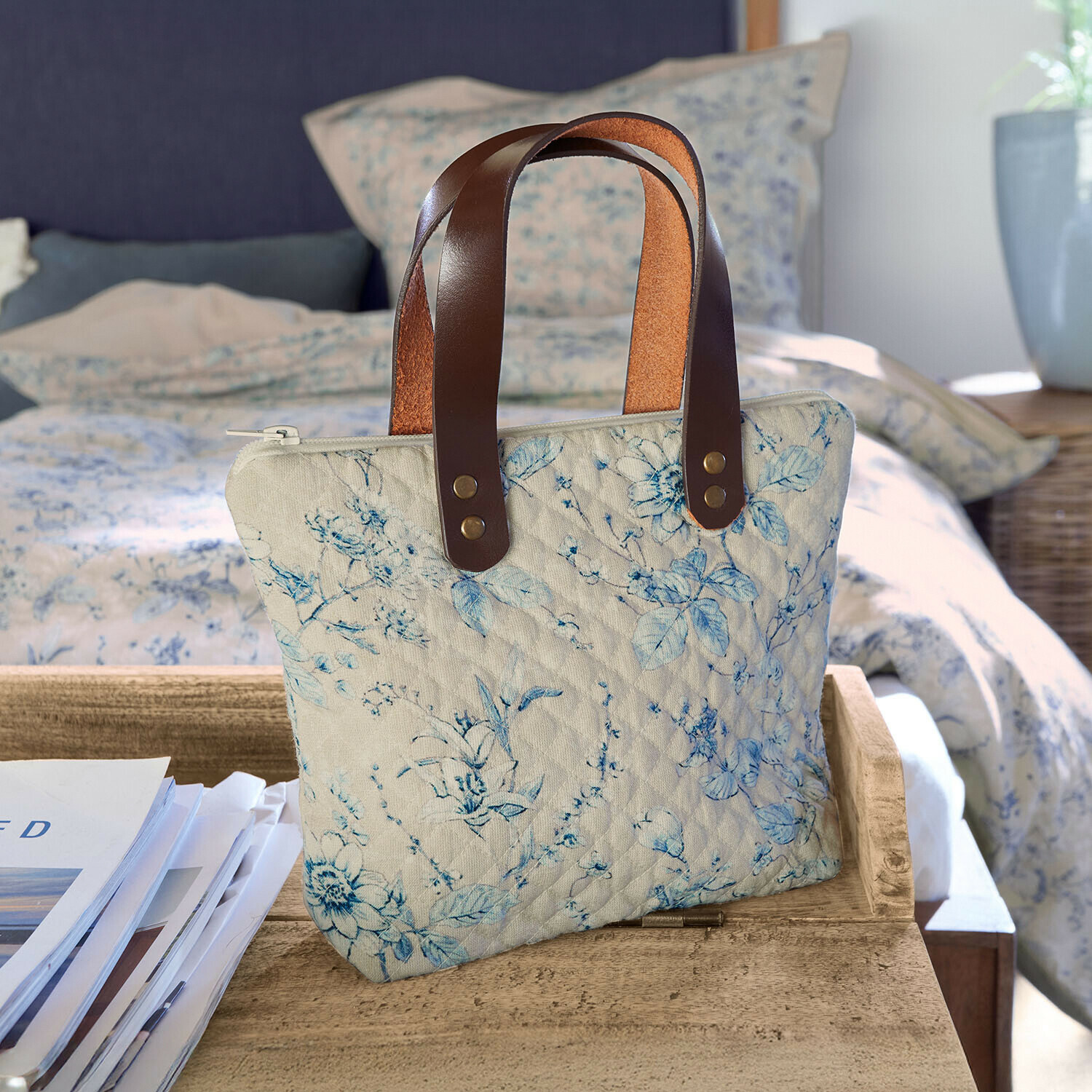Bolso acolchado beige con estampado floral azul y asas de cuero marrón sobre mesa de madera