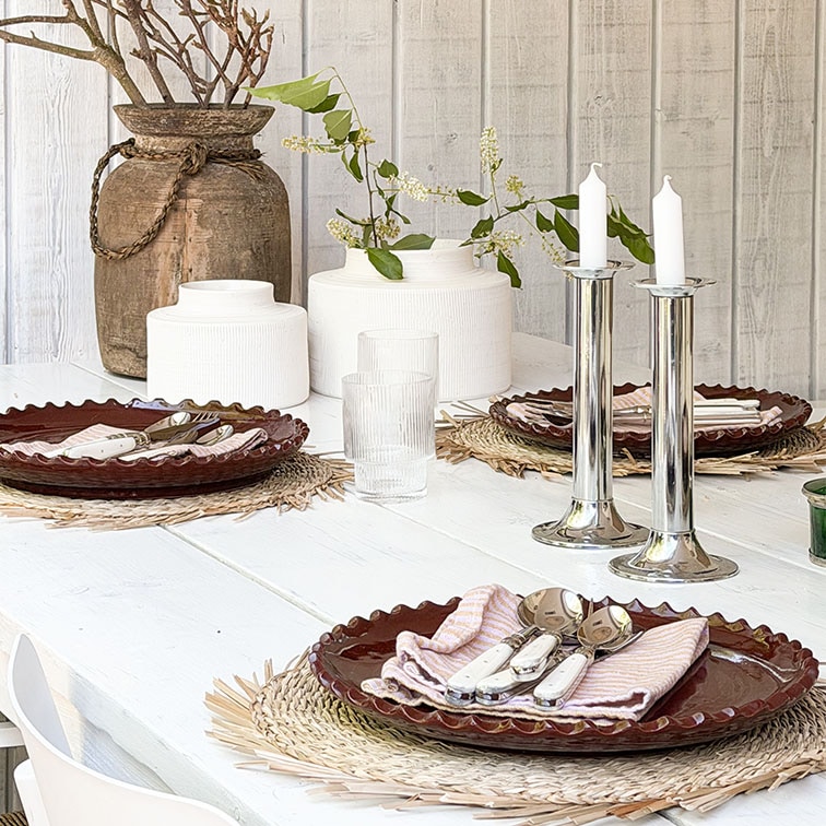 Mesa de madera blanca con platos de cerámica marrón y candelabros plateados sobre manteles decorativos