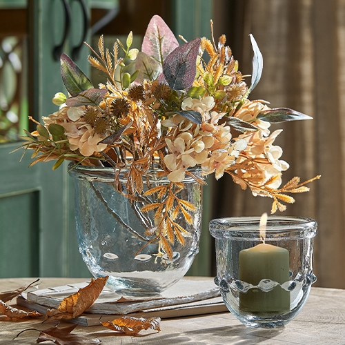 Jarrón de cristal con flores secas en tonos beige y marrón y portavelas con vela sobre mesa de madera