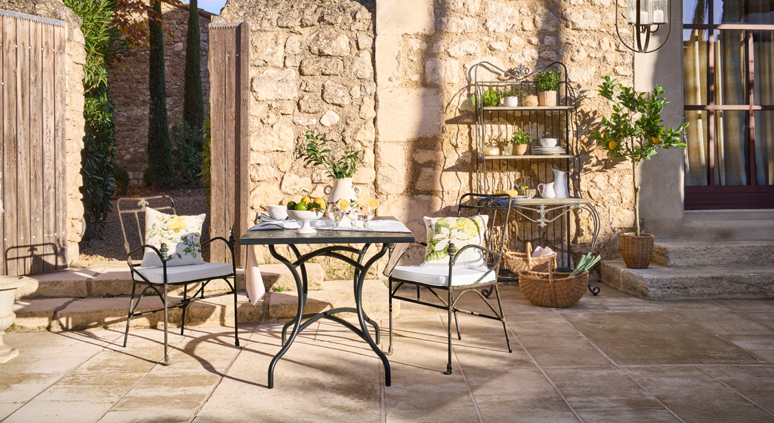 Mesa y dos sillas de metal en patio con pared de piedra y plantas, ambiente para desayuno