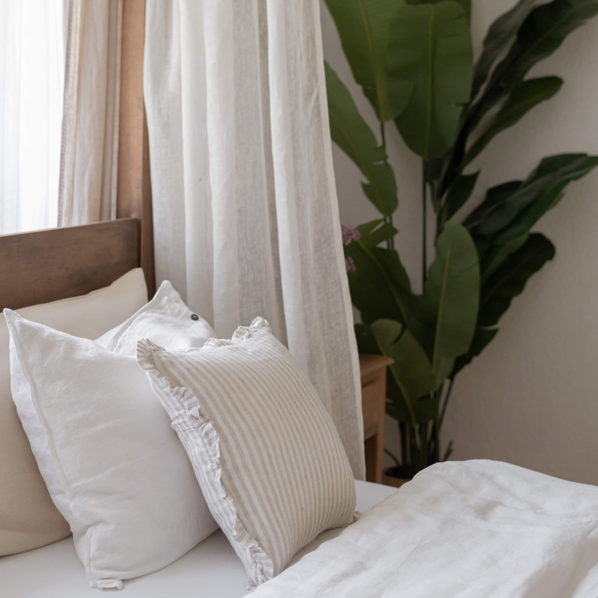 Cojines de lino blanco y beige en cama con cabecero de madera y planta verde al fondo