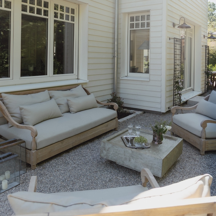 Conjunto de muebles de jardín de madera clara con cojines grises en patio de grava frente a casa blanca