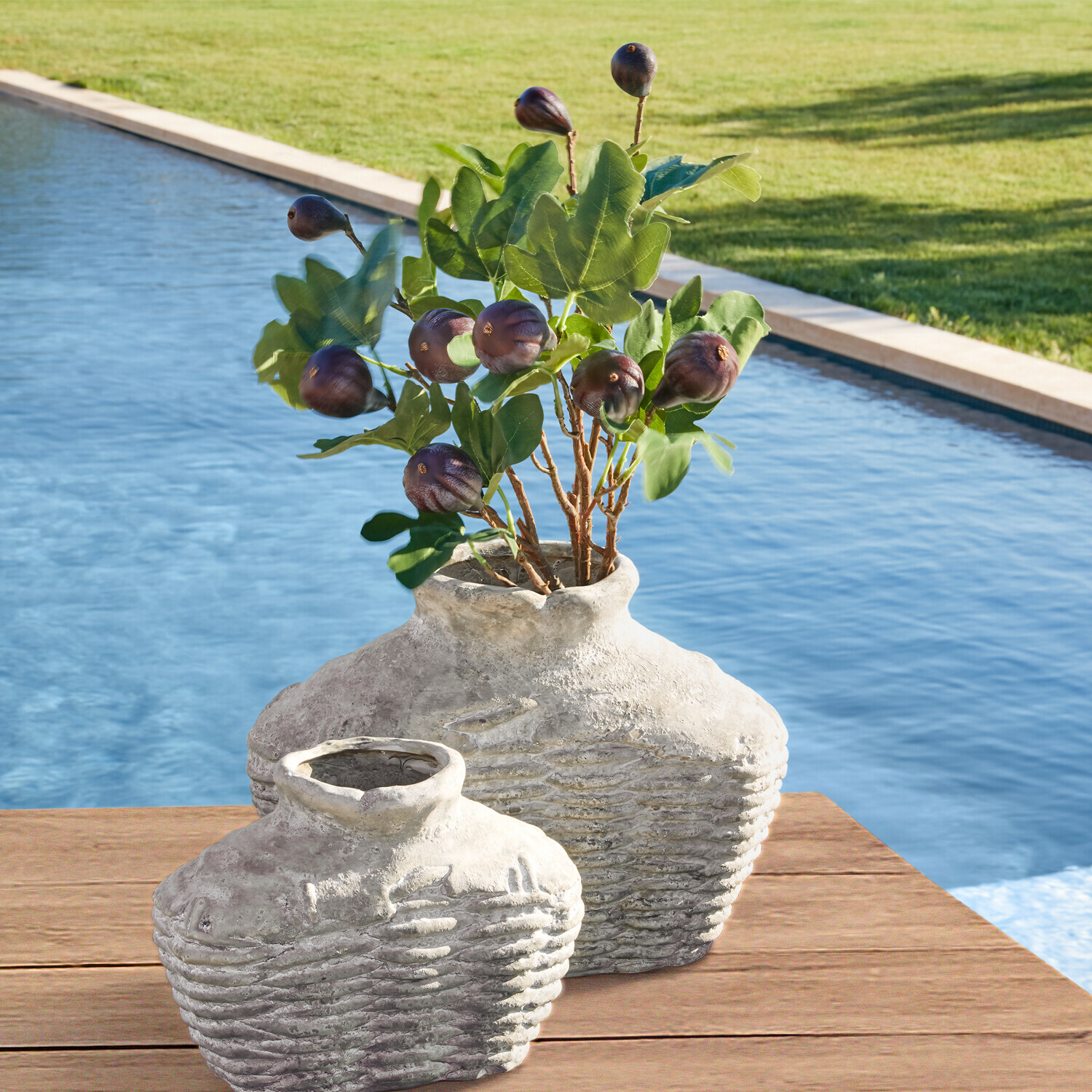 Dos jarrones rústicos de cerámica gris sobre mesa de madera junto a piscina con hojas verdes y frutos