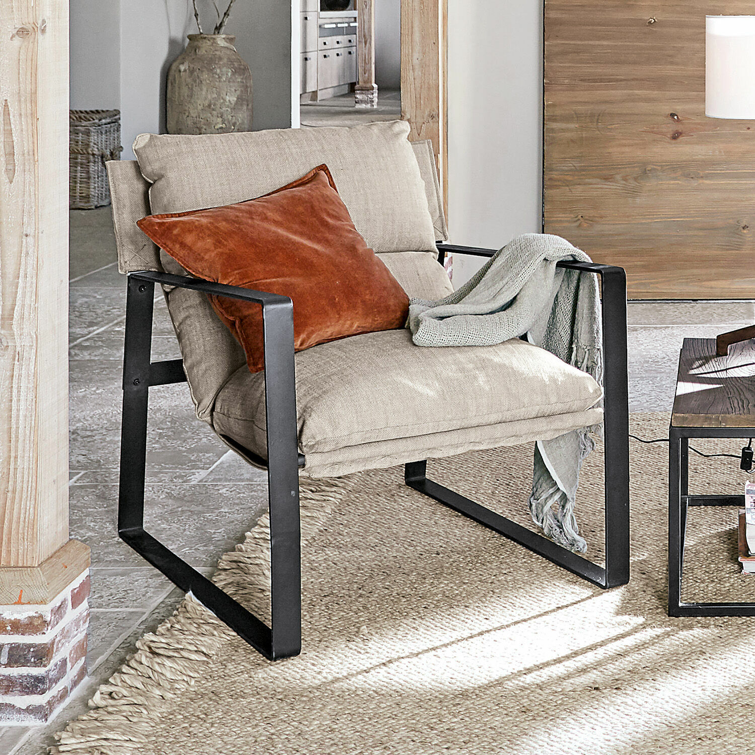 Sillón Seuvard Sillón de tela beige con estructura metálica negra, cojín naranja y manta gris en sala de estar