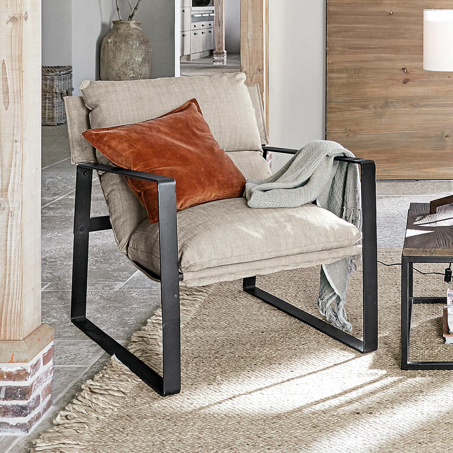 Sillón Seuvard Sillón de tela beige con estructura metálica negra, cojín naranja y manta gris en sala de estar