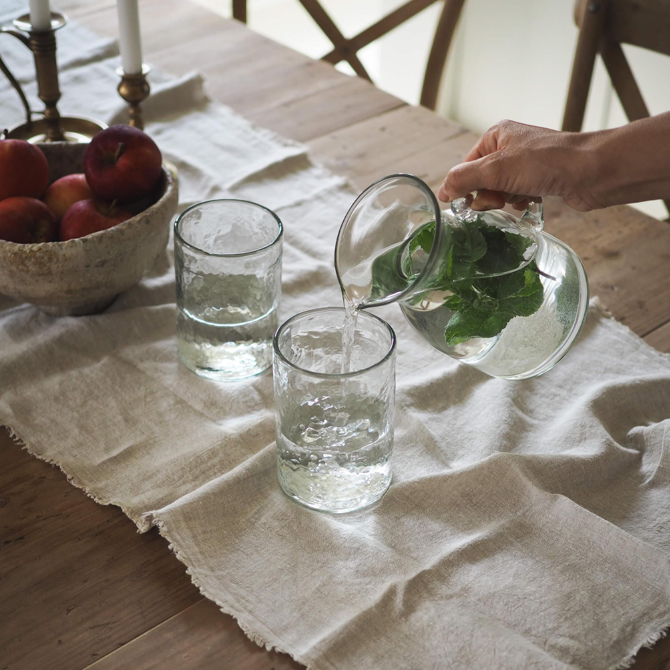 Jarra de vidrio con hojas verdes y dos vasos sobre un mantel de lino en mesa de madera