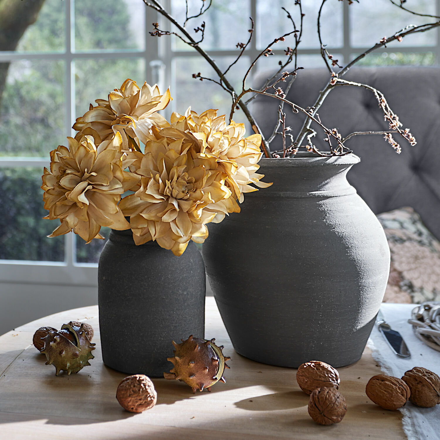 Set de 4 flores decorativas Cappellina Dos jarrones de cerámica gris oscuro con flores secas y nueces sobre mesa de madera clara
