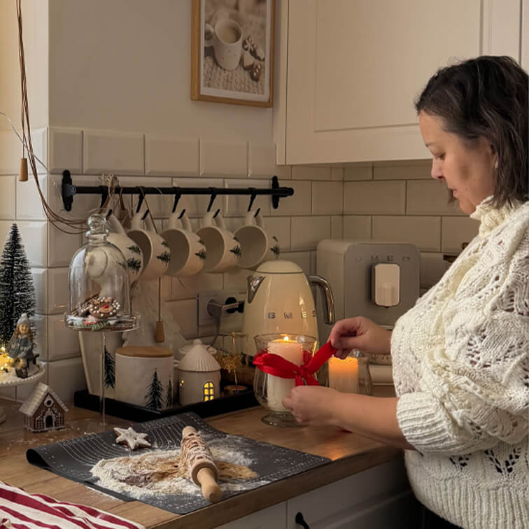 Mujer con suéter blanco decorando vela con lazo rojo en cocina acogedora con utensilios para hornear