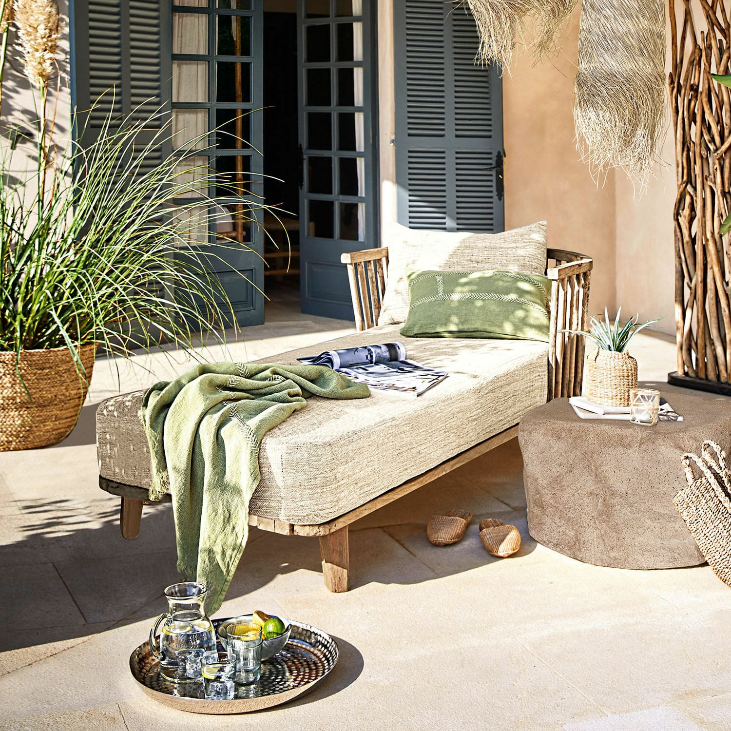 Chaise longue Bellville Tumbona de madera con cojín beige y manta verde en terraza decorada con bandeja de bebidas