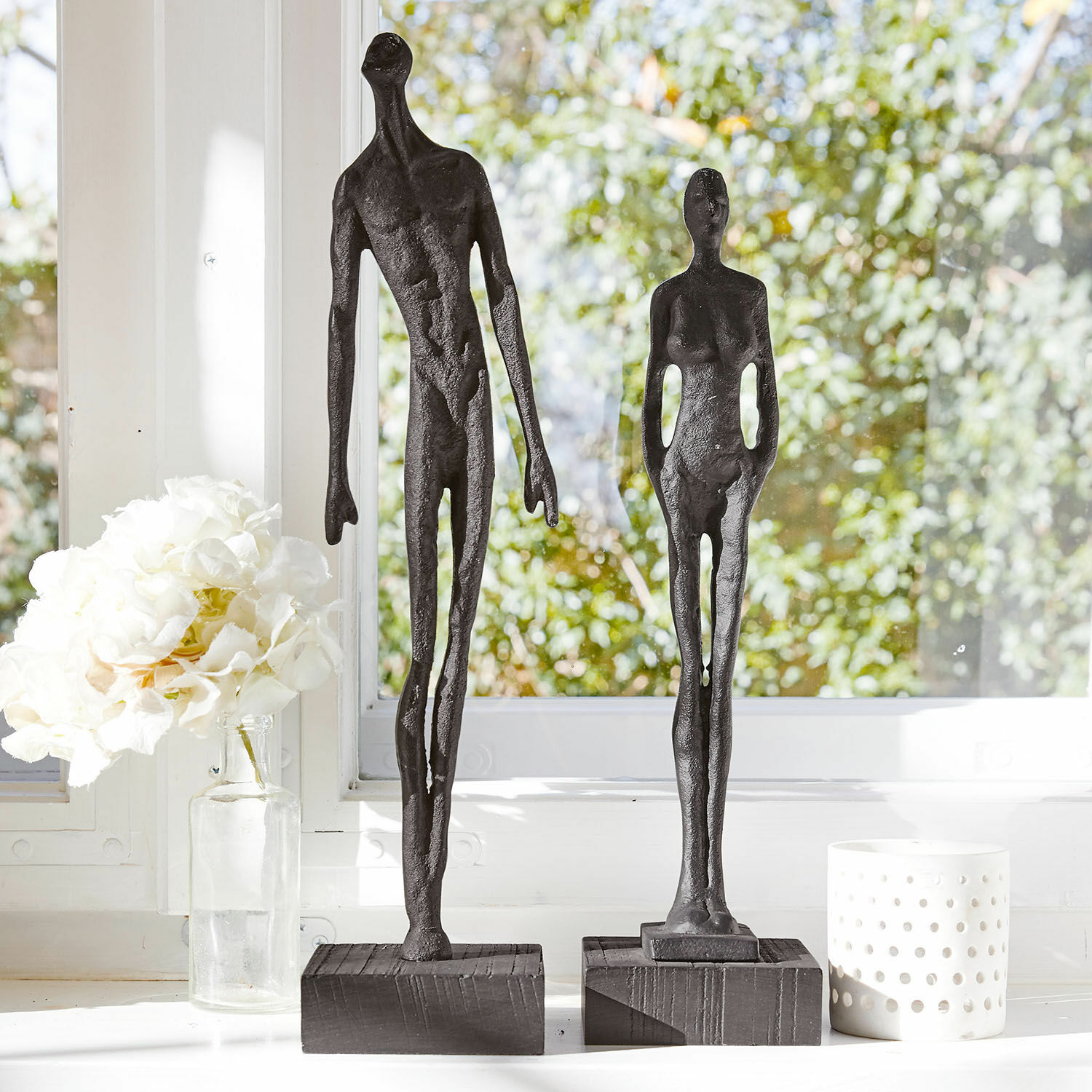 Set de 2 figuras Perce Dos esculturas negras de figuras humanas en metal sobre bases de madera frente a ventana luminosa