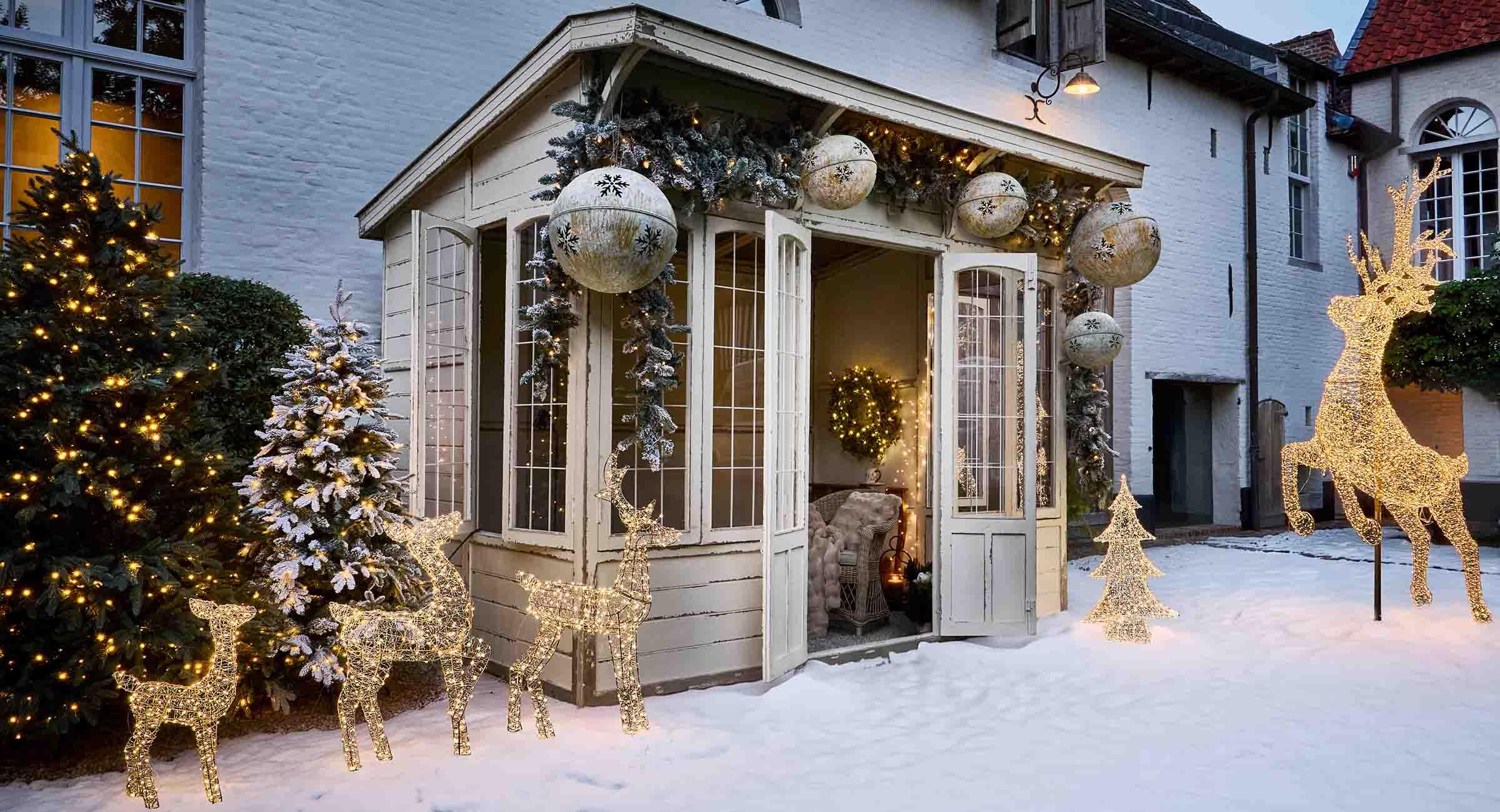 Decoraciones luminosas de renos y árboles de Navidad en la nieve frente a casa blanca con ambiente invernal