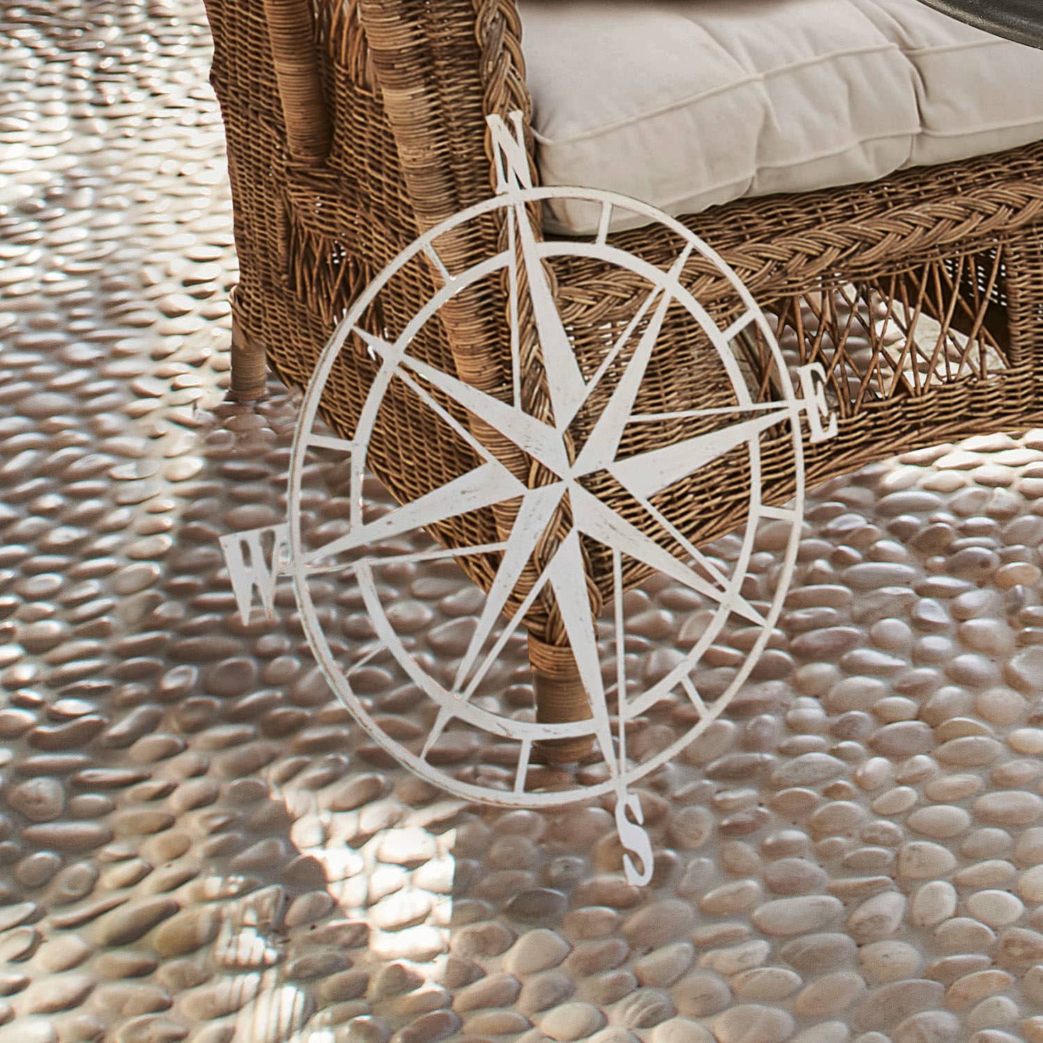 Rosa de los vientos Bianco Rosa de los vientos decorativa de metal blanco frente a sillón de mimbre sobre suelo de piedras