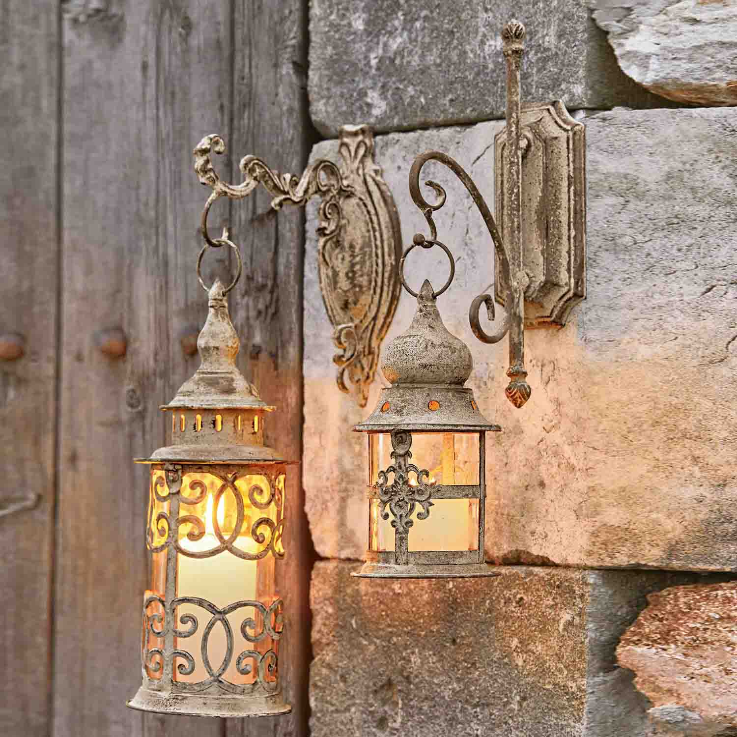 Dos faroles de pared rústicos de metal en color beige envejecido iluminando una pared de piedra