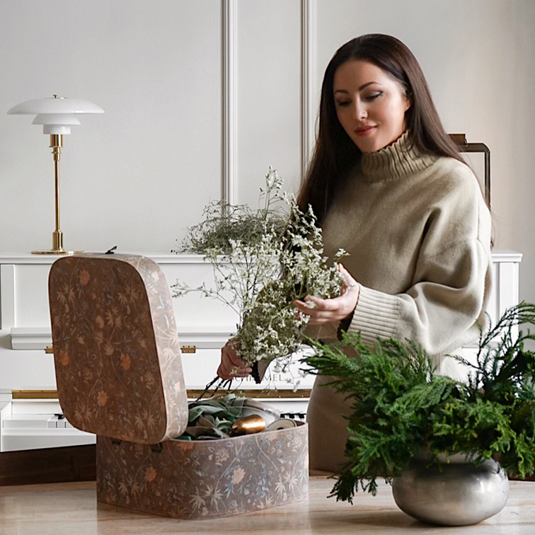 Mujer con suéter beige sosteniendo ramo de flores junto a caja decorativa y planta verde sobre mesa