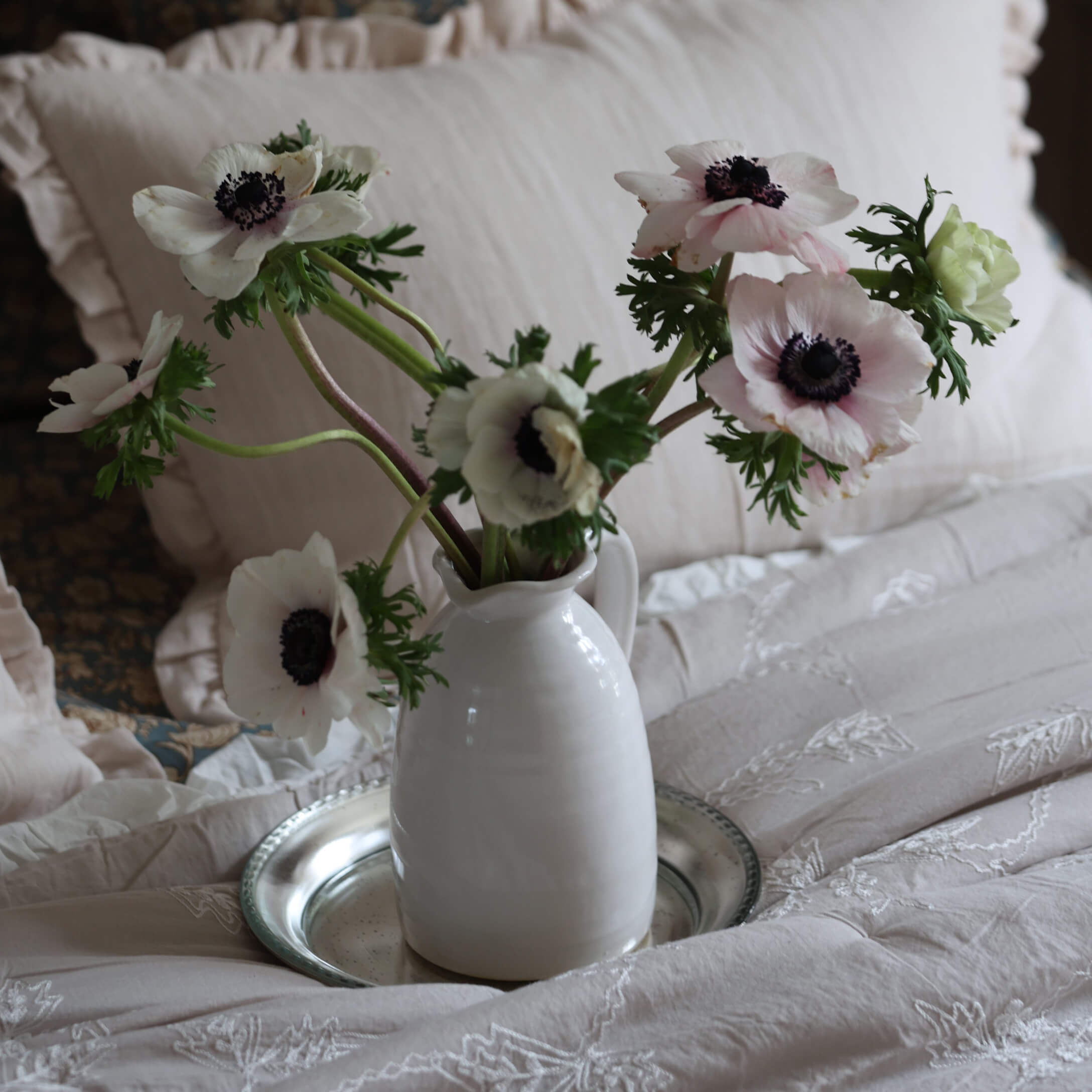 Jarra de cerámica blanca con delicadas anémonas blancas sobre ropa de cama beige bordada