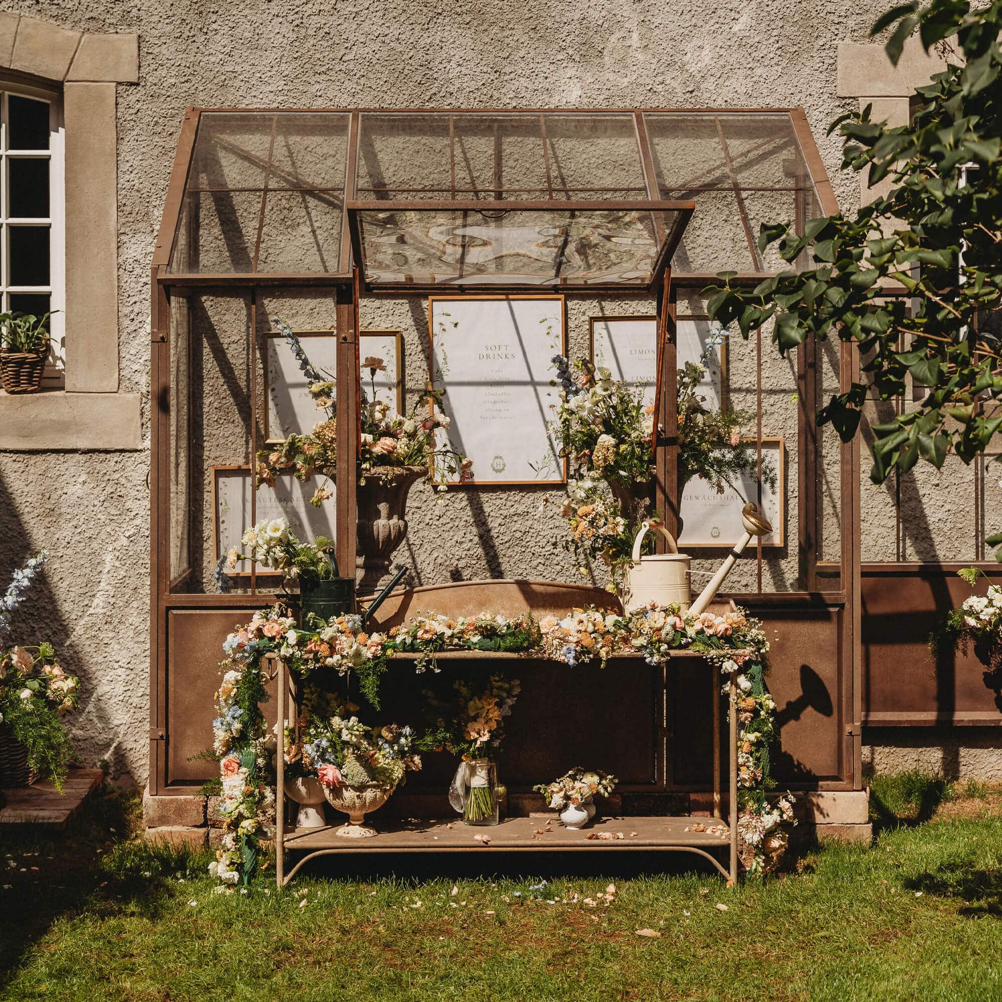 Mesa de jardín de metal con flores y regaderas frente a un invernadero de cristal