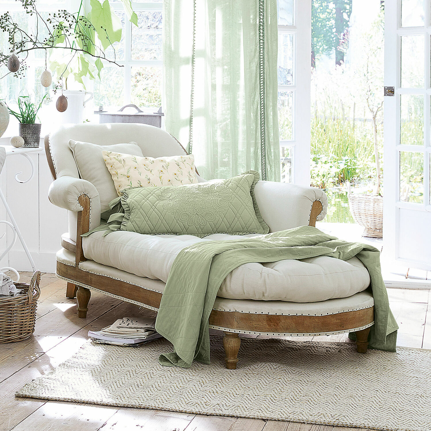 Chaise longue Queyranne Chaiselongue elegante de madera clara y tela crema con manta verde y cojines en interior luminoso