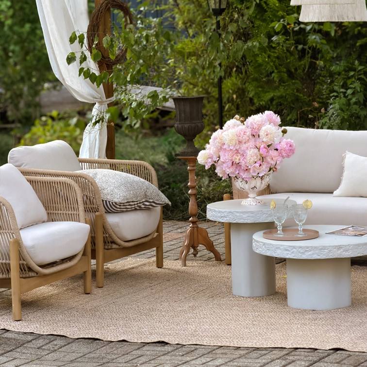 Conjunto de muebles de jardín con cojines claros, sillones de mimbre y mesas de cemento sobre alfombra de yute