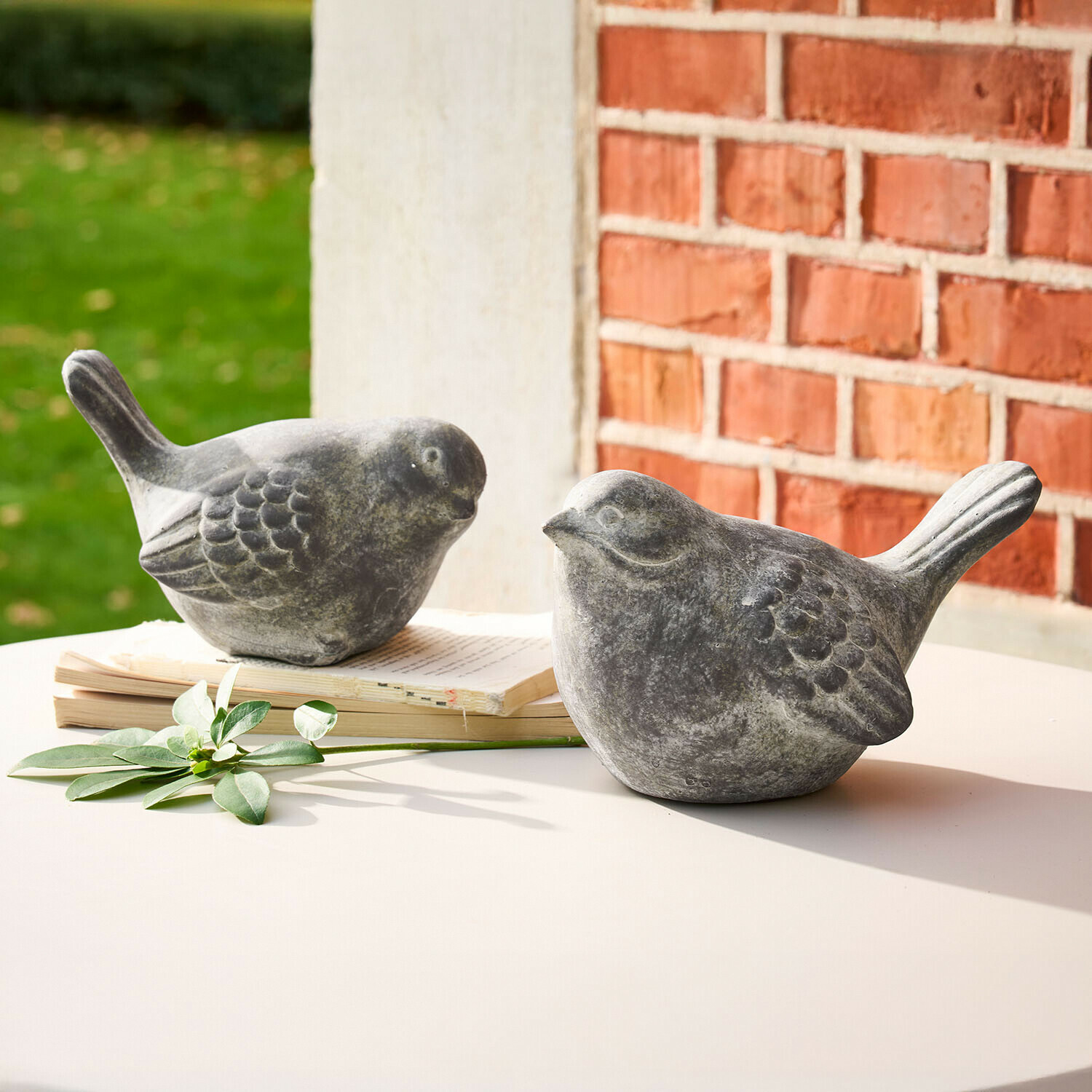 Dos figuras decorativas de pájaros en piedra gris sobre mesa blanca con libro y hoja verde