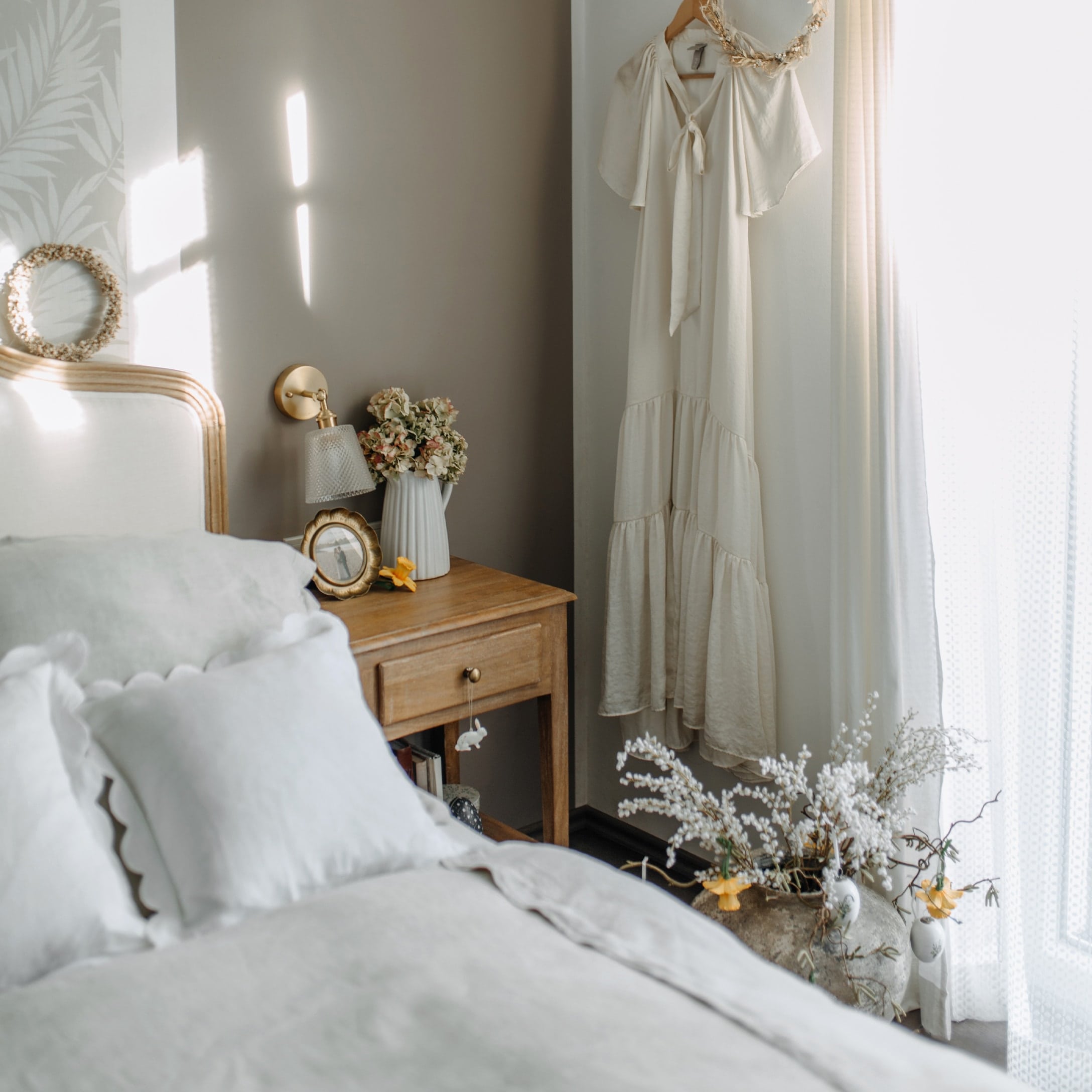 Dormitorio acogedor con mesita de noche de madera, ropa de cama blanca y vestido crema colgado en la pared