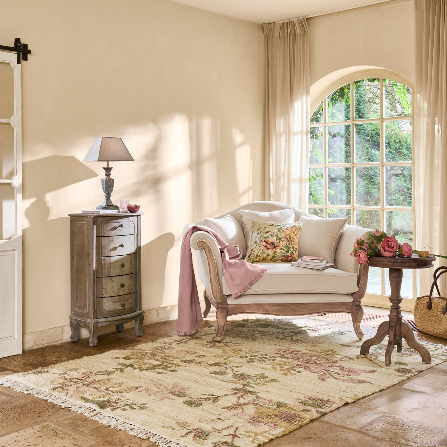 Sala acogedora con sofá beige de madera, cojín floral y alfombra de tonos pastel junto a ventana grande