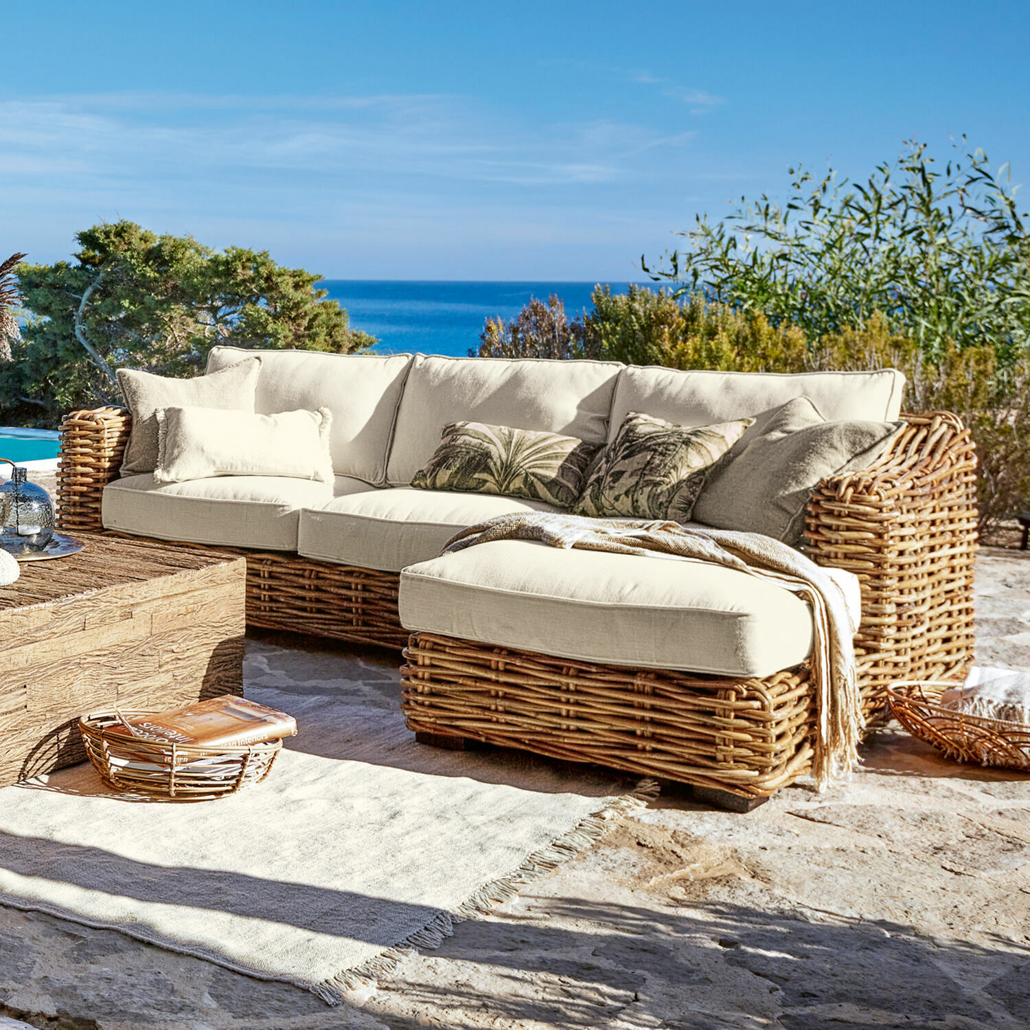 Sofá Villedieu Sofá esquinero de ratán con cojines crema en terraza con vista al mar, muebles de jardín