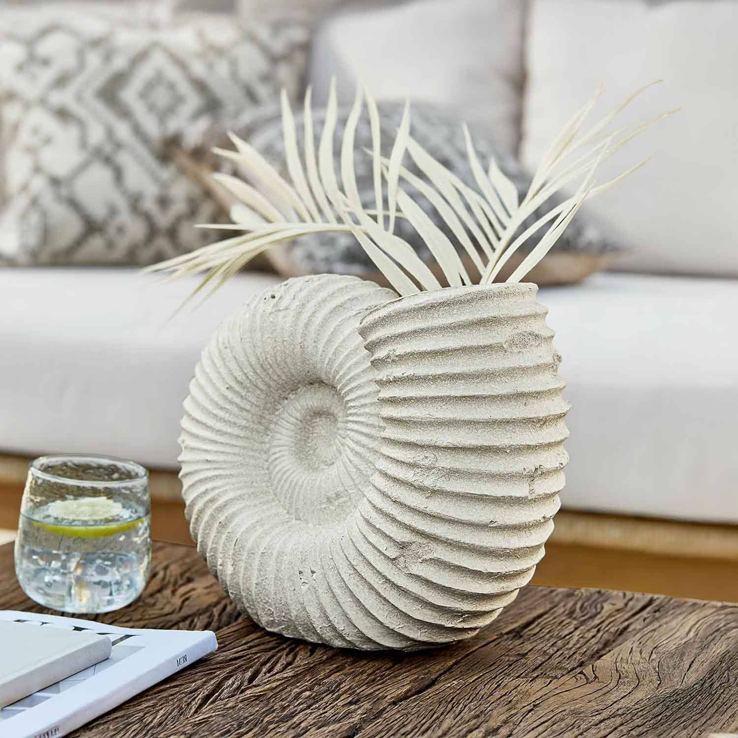 Jarrón decorativo de piedra clara con forma de concha y hojas secas sobre mesa de madera