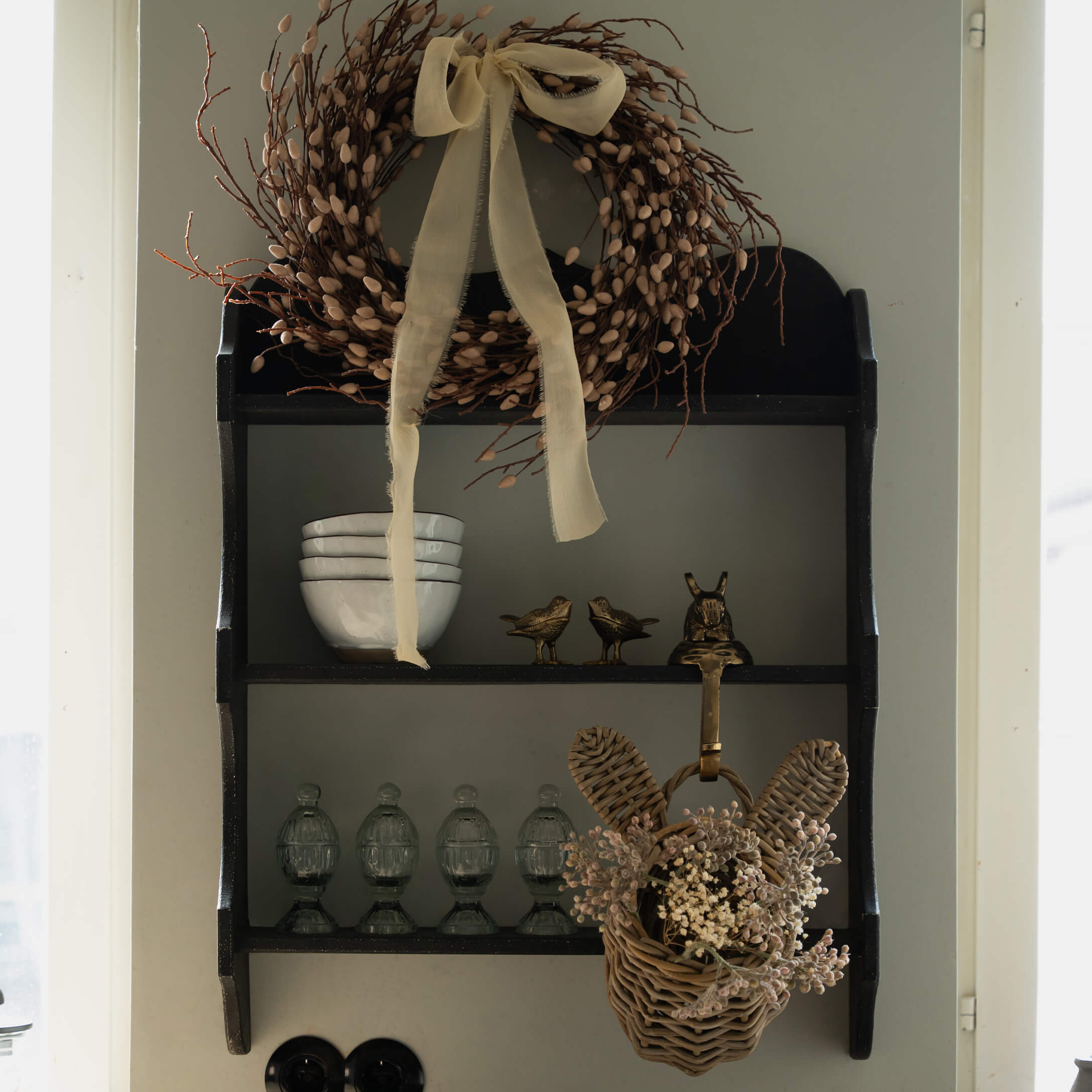 Estantería de madera negra con corona decorativa y cesta de mimbre con flores secas colgada