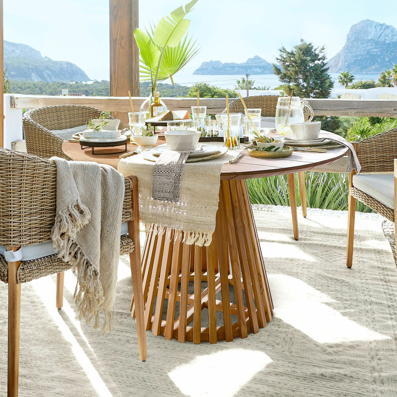 Mesa redonda de madera con decoración y sillas de mimbre en terraza con vistas a montañas y mar