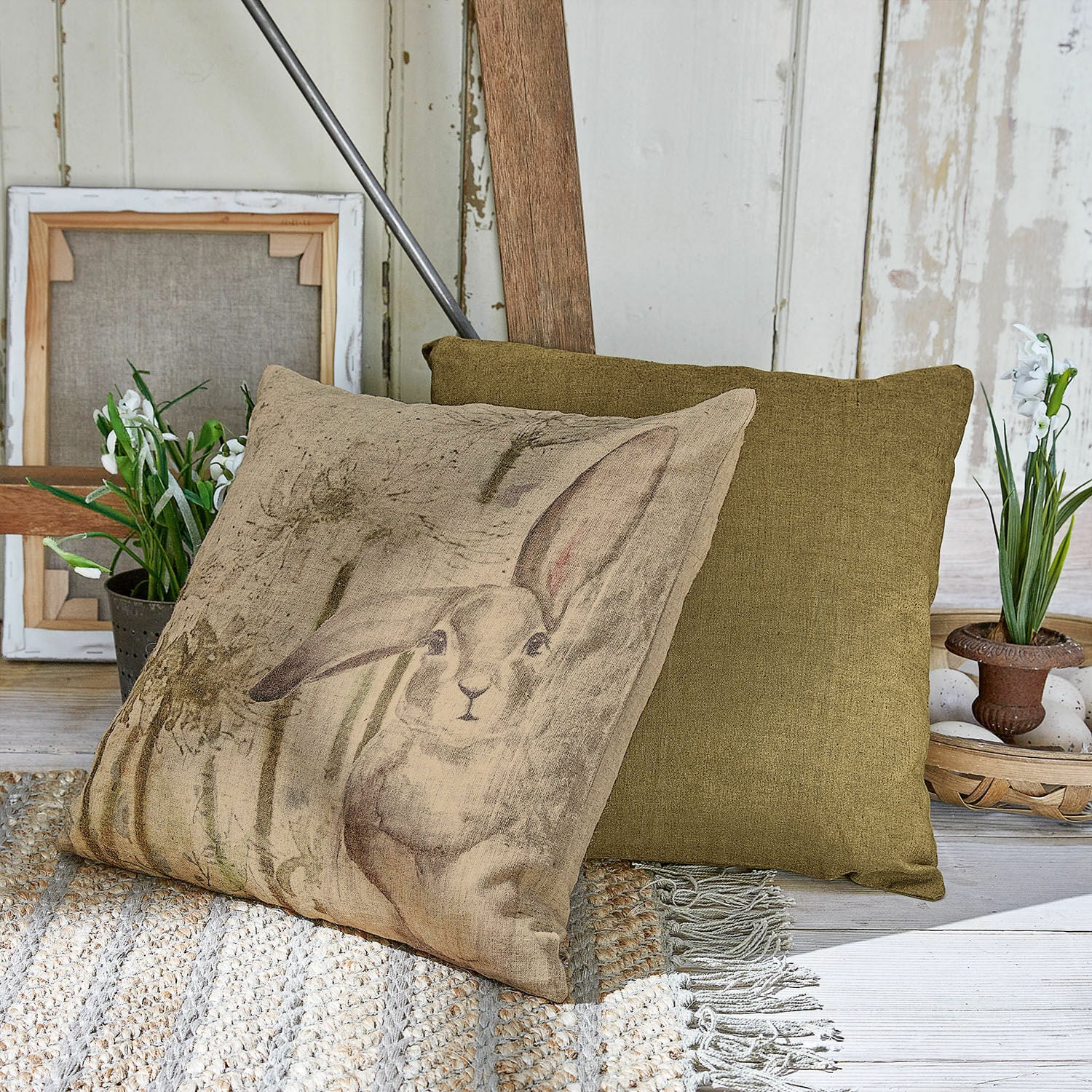 Set de 2 fundas de cojín Milda Cojín beige con estampado de conejo y cojín verde decorativo sobre alfombra tejida
