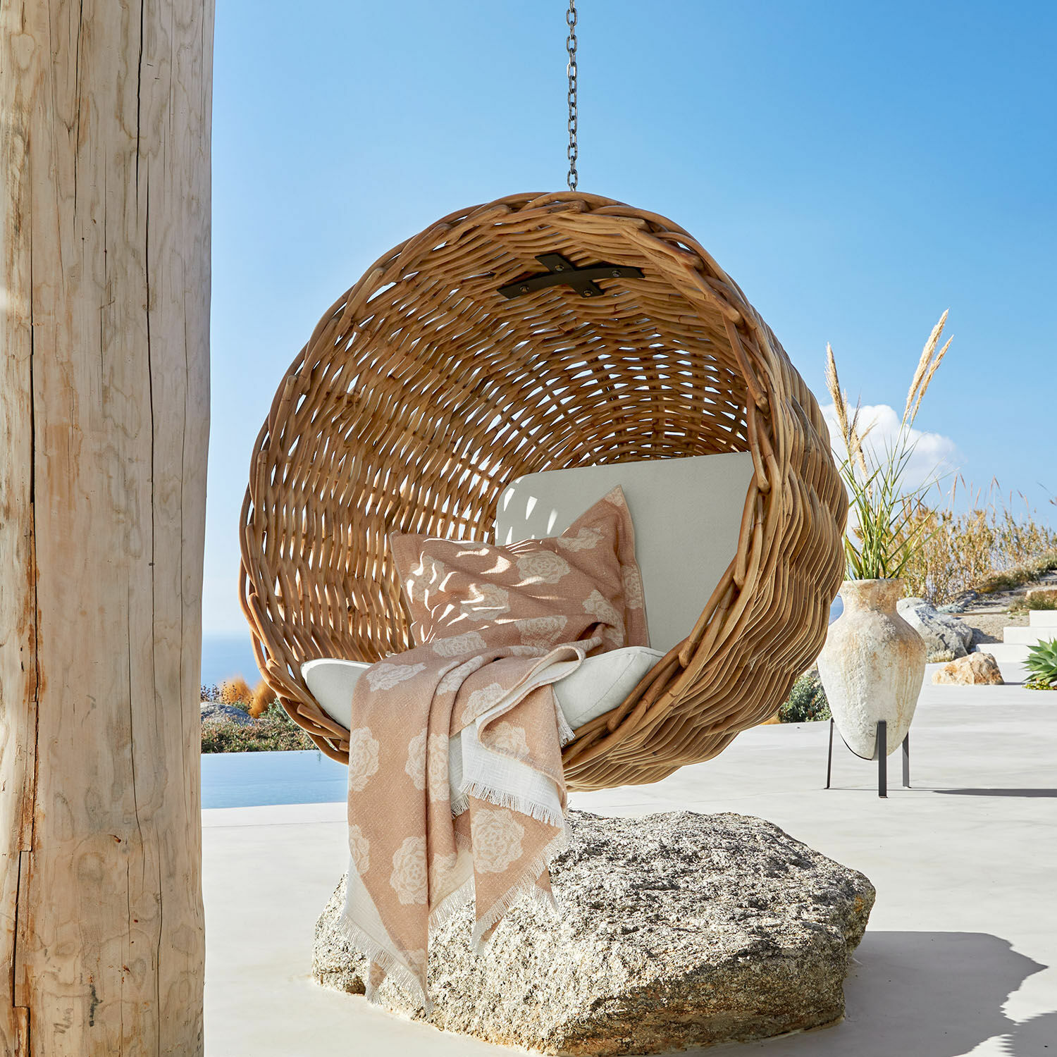 Silla colgante Nirevox Sillón colgante de ratán natural con cojín claro y manta rosa en terraza con vista al mar