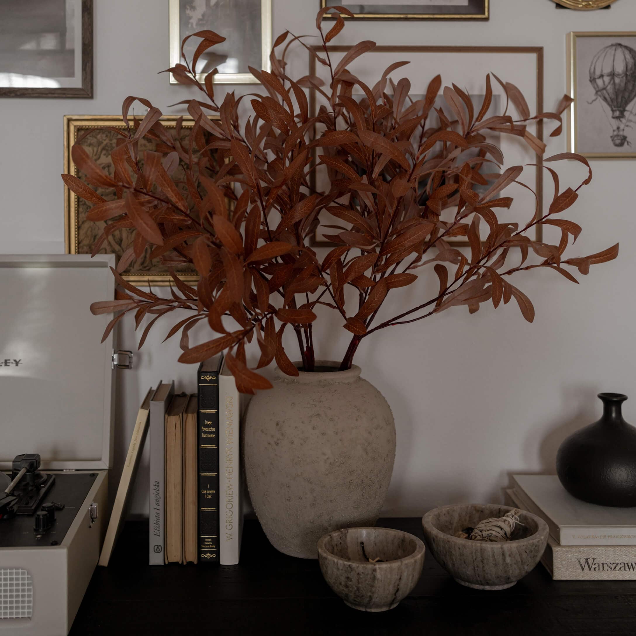 Jarrón de alabastro beige con ramas secas marrones sobre mesa oscura con libros y decoración