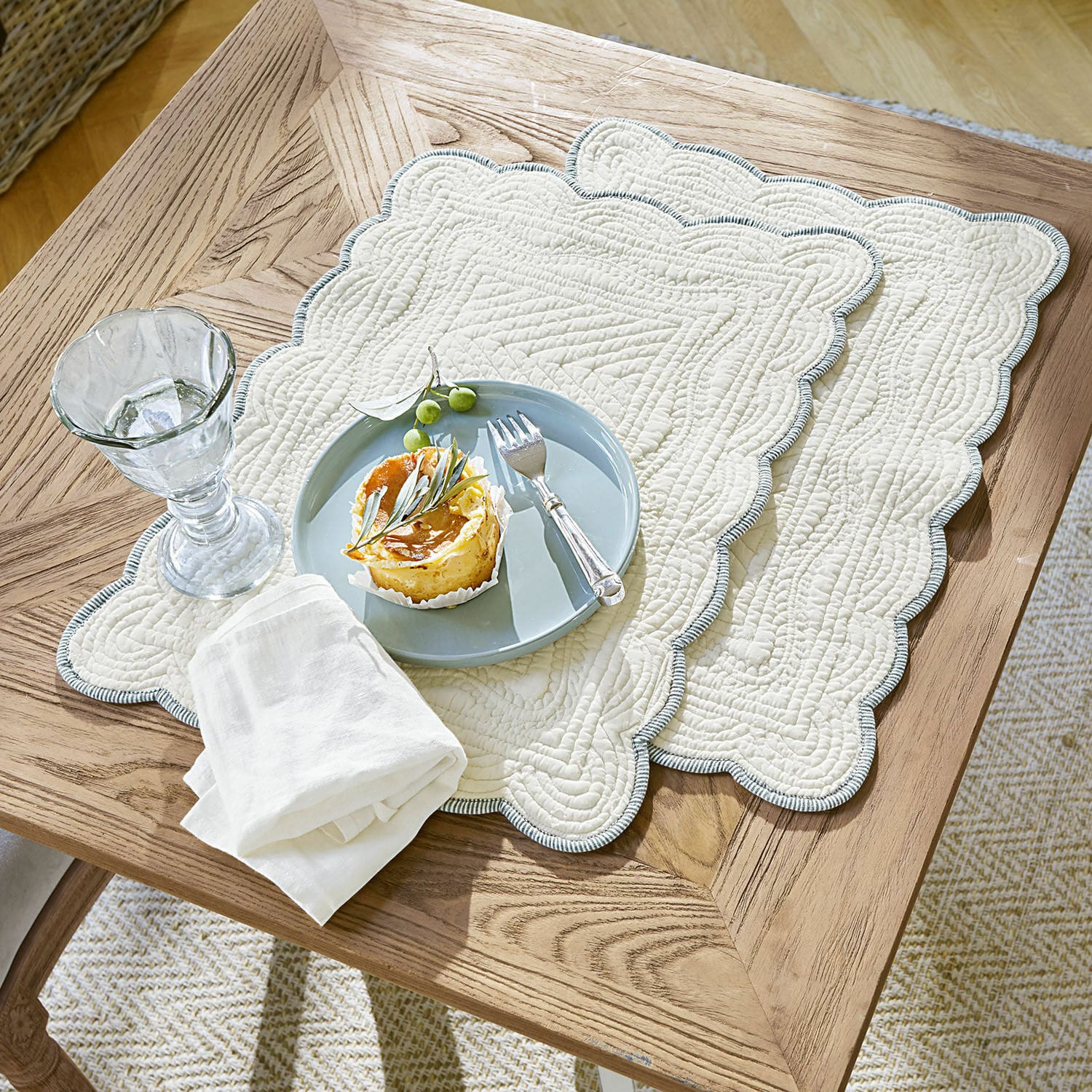 Juego de 2 manteles individuales Fontienne Individuales acolchados de algodón beige con borde azul sobre mesa de madera con postre y vaso
