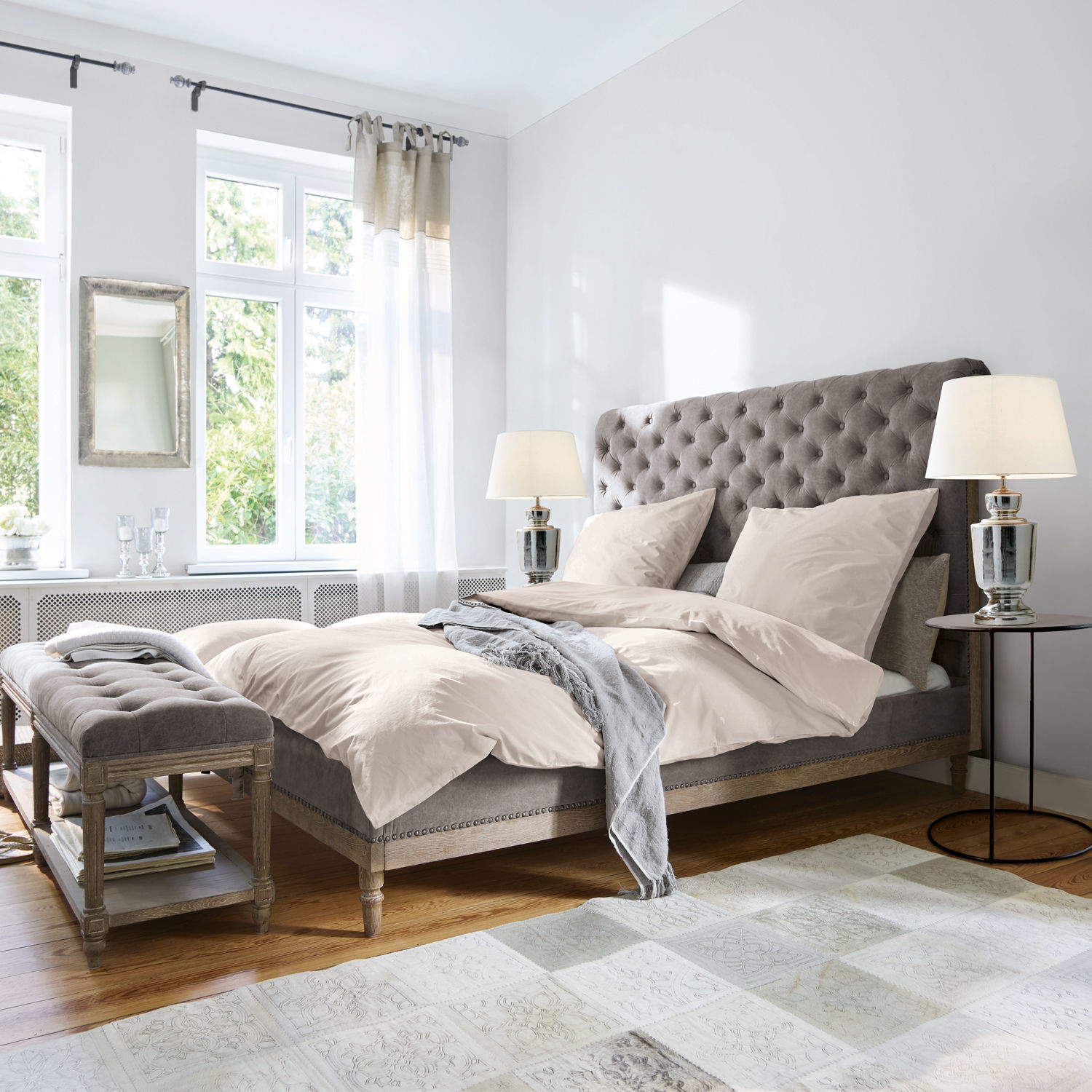 Cama Pozières Cama tapizada gris con cabecero capitoné y ropa de cama beige en dormitorio luminoso con suelo de madera