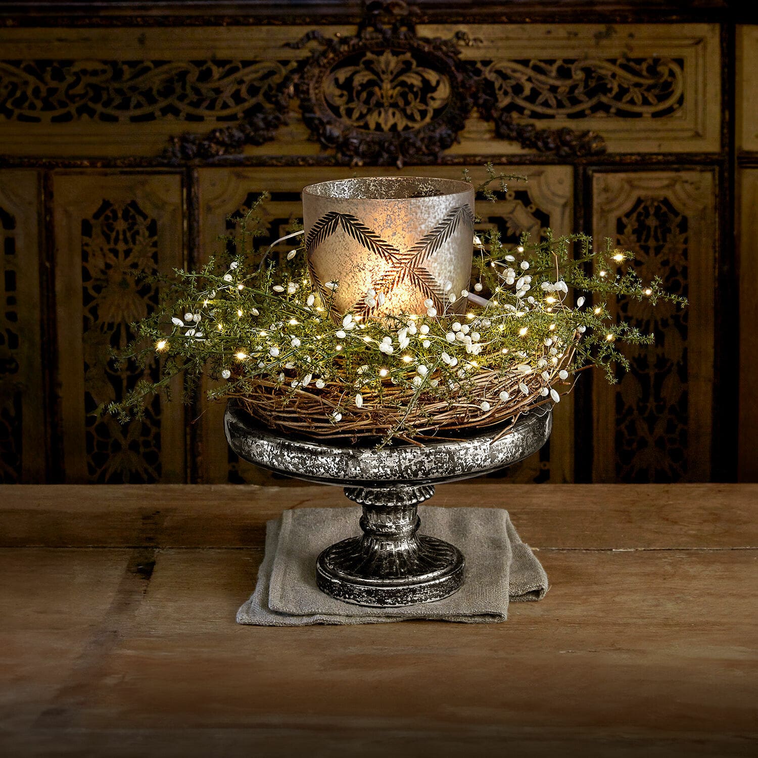 Bandeja con pie Incantesse Portavelas decorativo de metal plateado con corona de mimbre y luces LED sobre mesa de madera