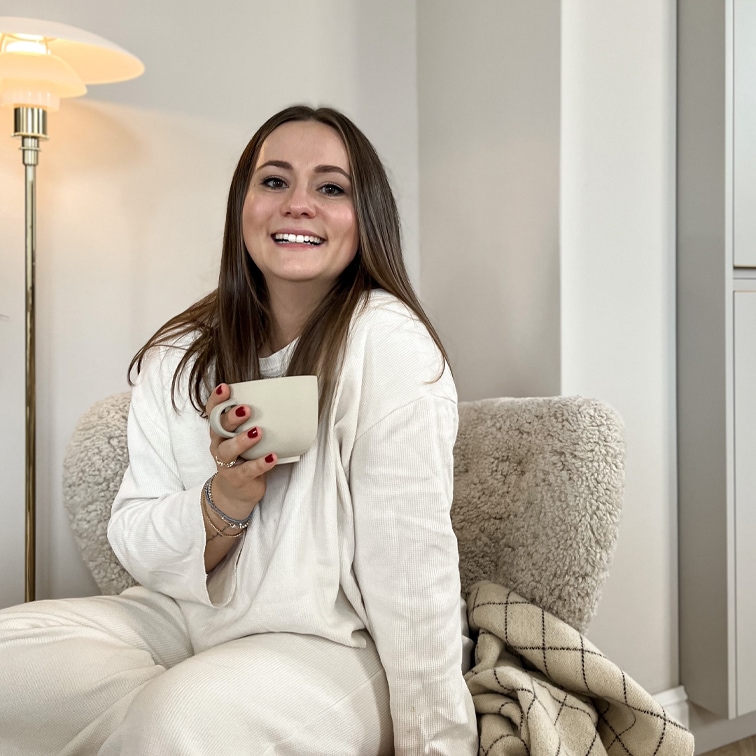 Mujer con ropa blanca suave sentada en sillón crema con taza, ambiente acogedor y cómodo