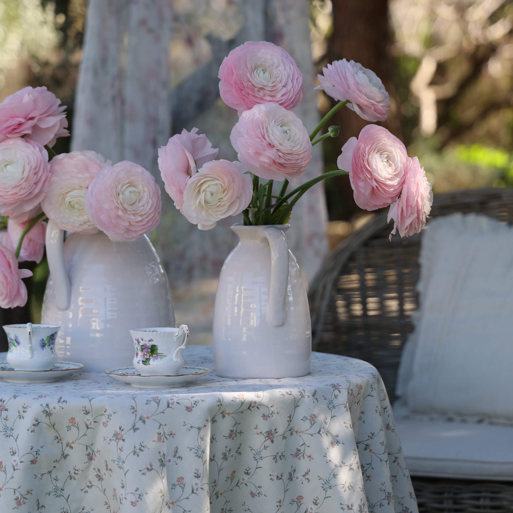 Jarras de cerámica blanca con flores rosas sobre mesa con mantel floral y tazas de té decorativas