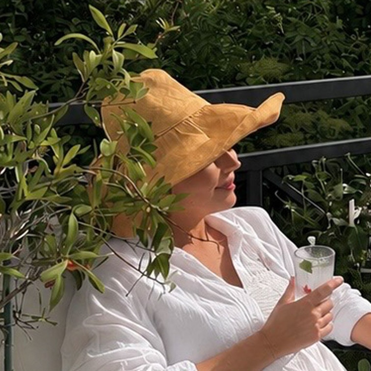 Mujer con blusa blanca y sombrero de paja amarillo descansando en terraza con bebida en mano