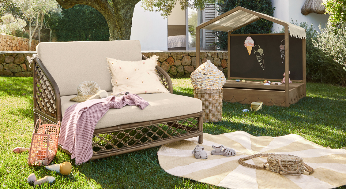 Jardín con sofá beige de ratán y casita de madera para niños sobre césped verde