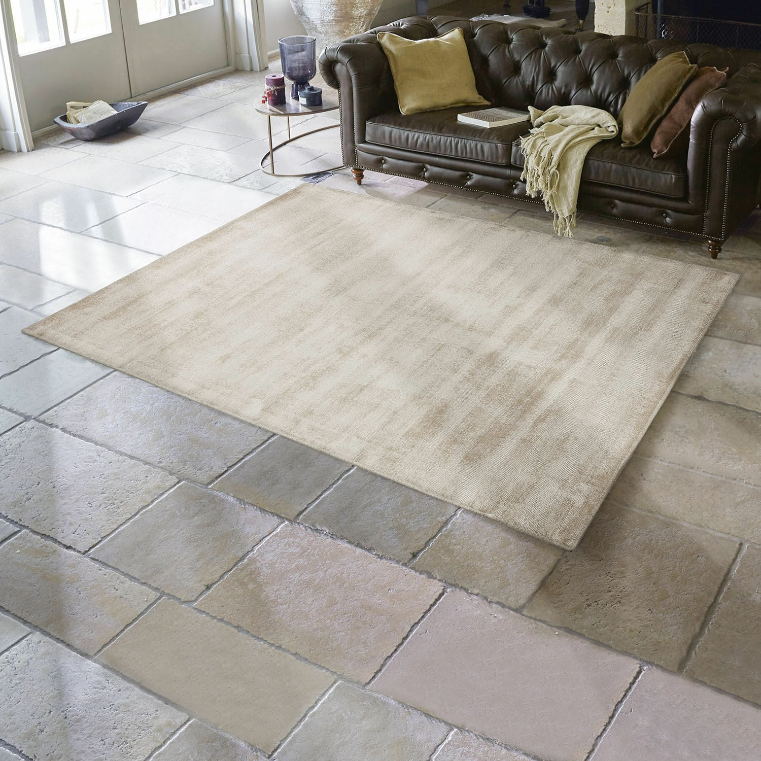 Alfombra Mariselle Alfombra beige de material suave sobre suelo de piedra en sala con sofá de cuero marrón oscuro