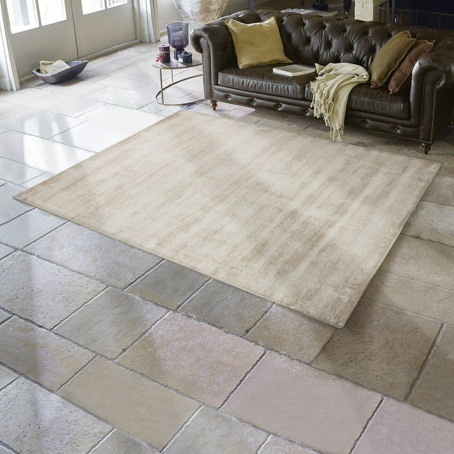 Alfombra Mariselle Alfombra beige de material suave sobre suelo de piedra en sala con sofá de cuero marrón oscuro