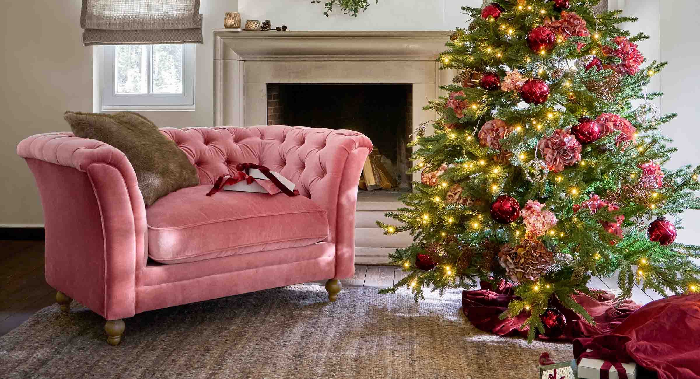 Sofá de terciopelo rosa junto a un árbol de Navidad decorado con bolas rojas y luces festivas