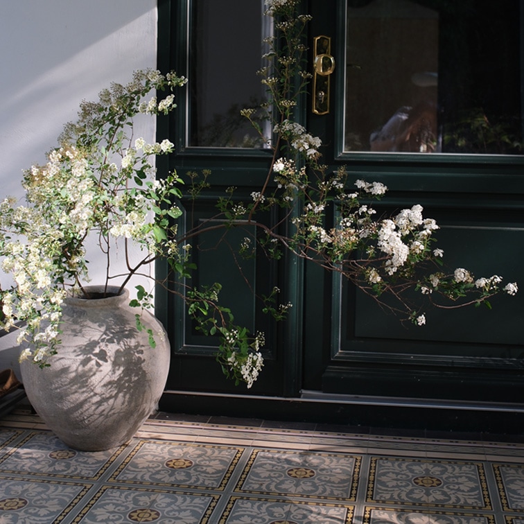 Jarrón grande de cerámica con flores blancas frente a puerta oscura y suelo de baldosas decorativas